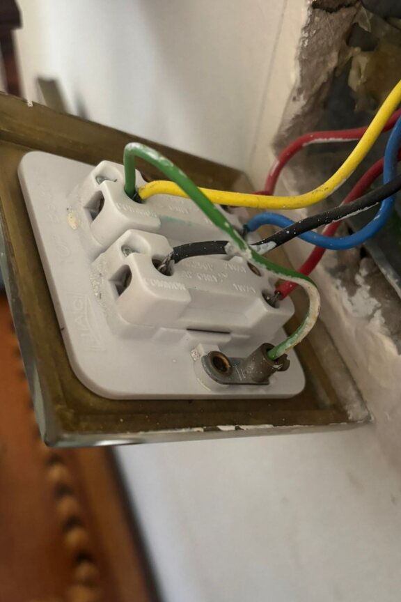 Rear of a metal light switch showing a green-and-yellow conductor incorrectly connected between a permanent live terminal and the metal plate earth terminal